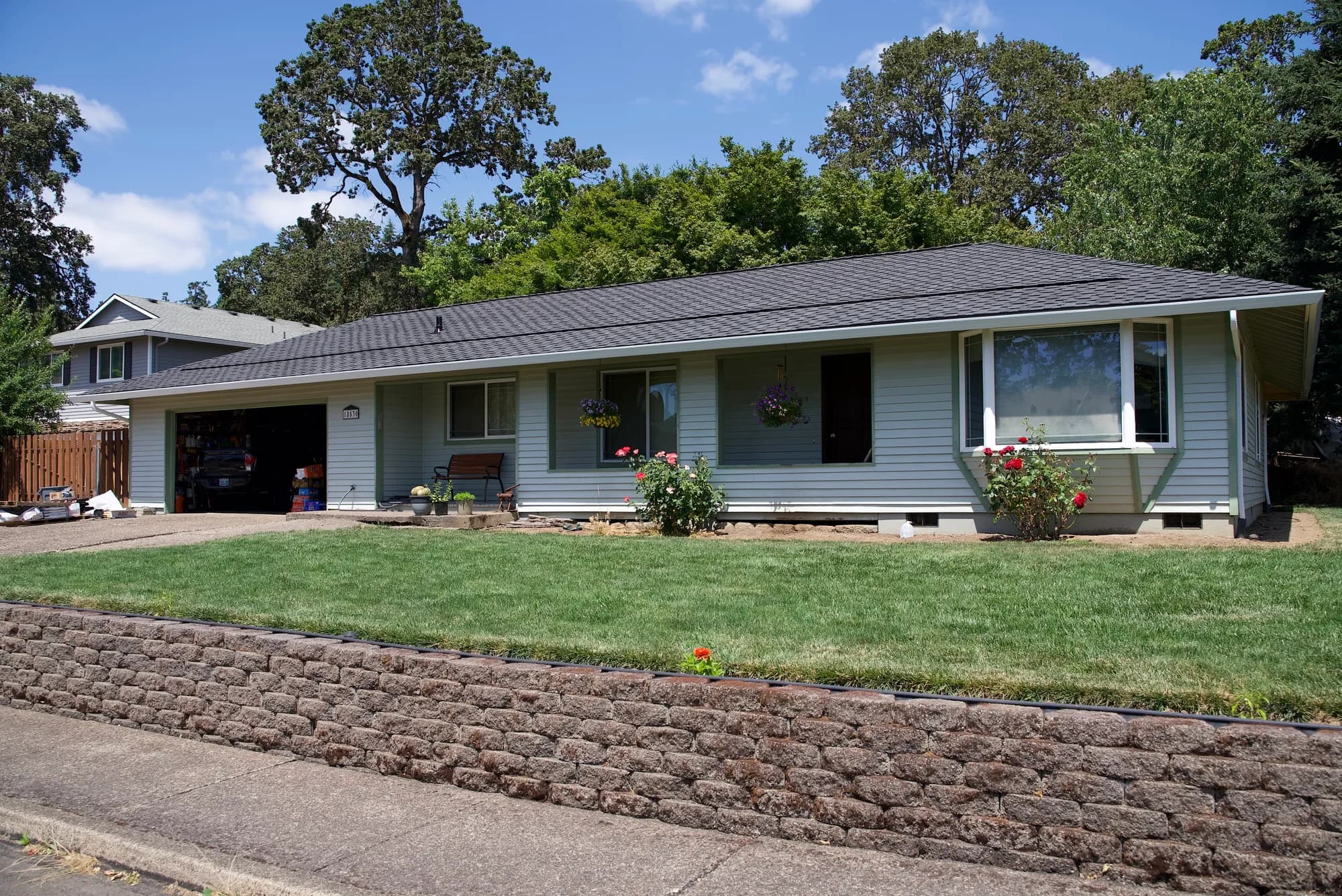Front elevation of a ranch house with a new dark gray shingle roof, bay window, and rose bushes in Gladstone, OR
