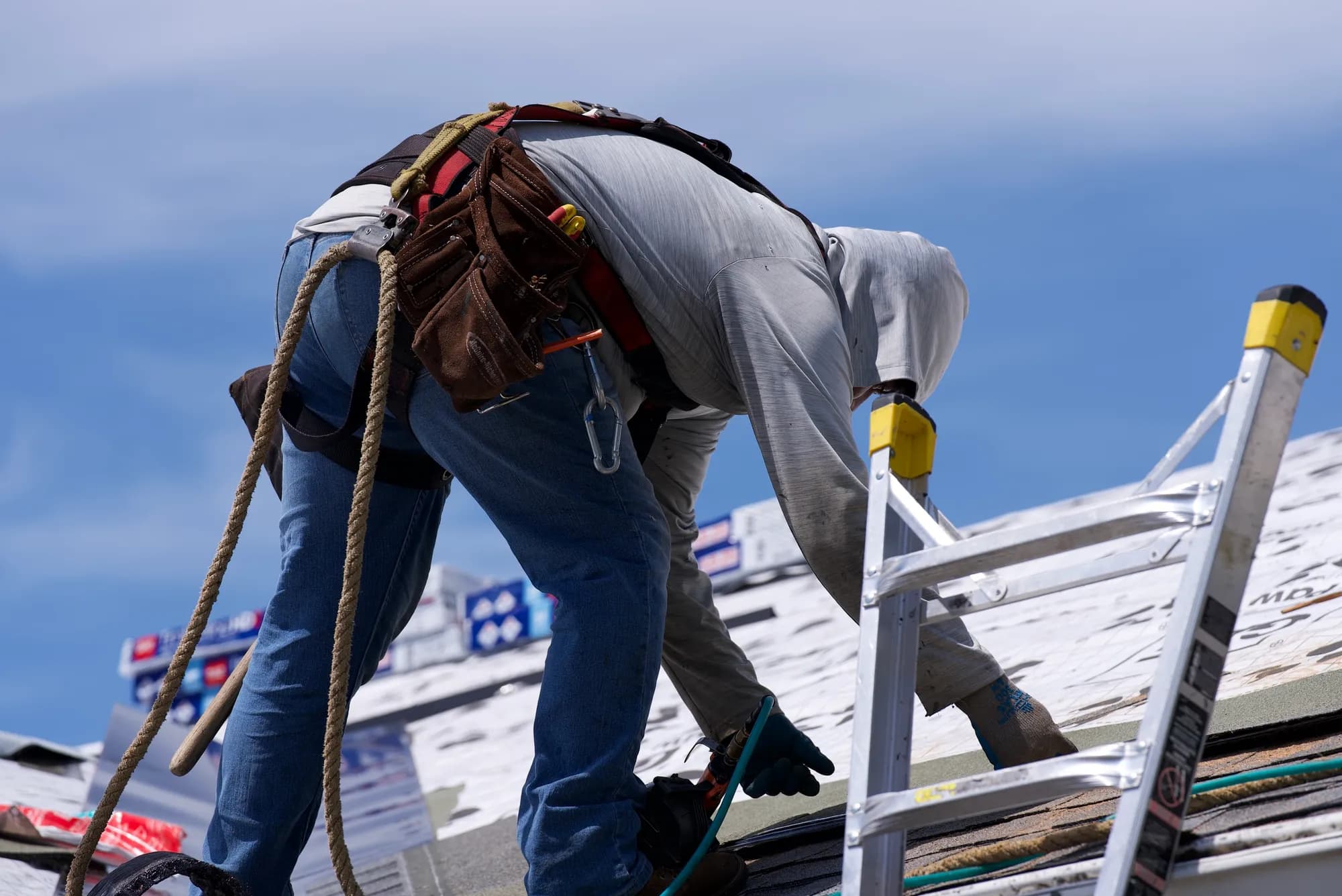 A harnessed roofer with a leather tool belt bending over printed underlayment next to a ladder in Hillsboro, OR