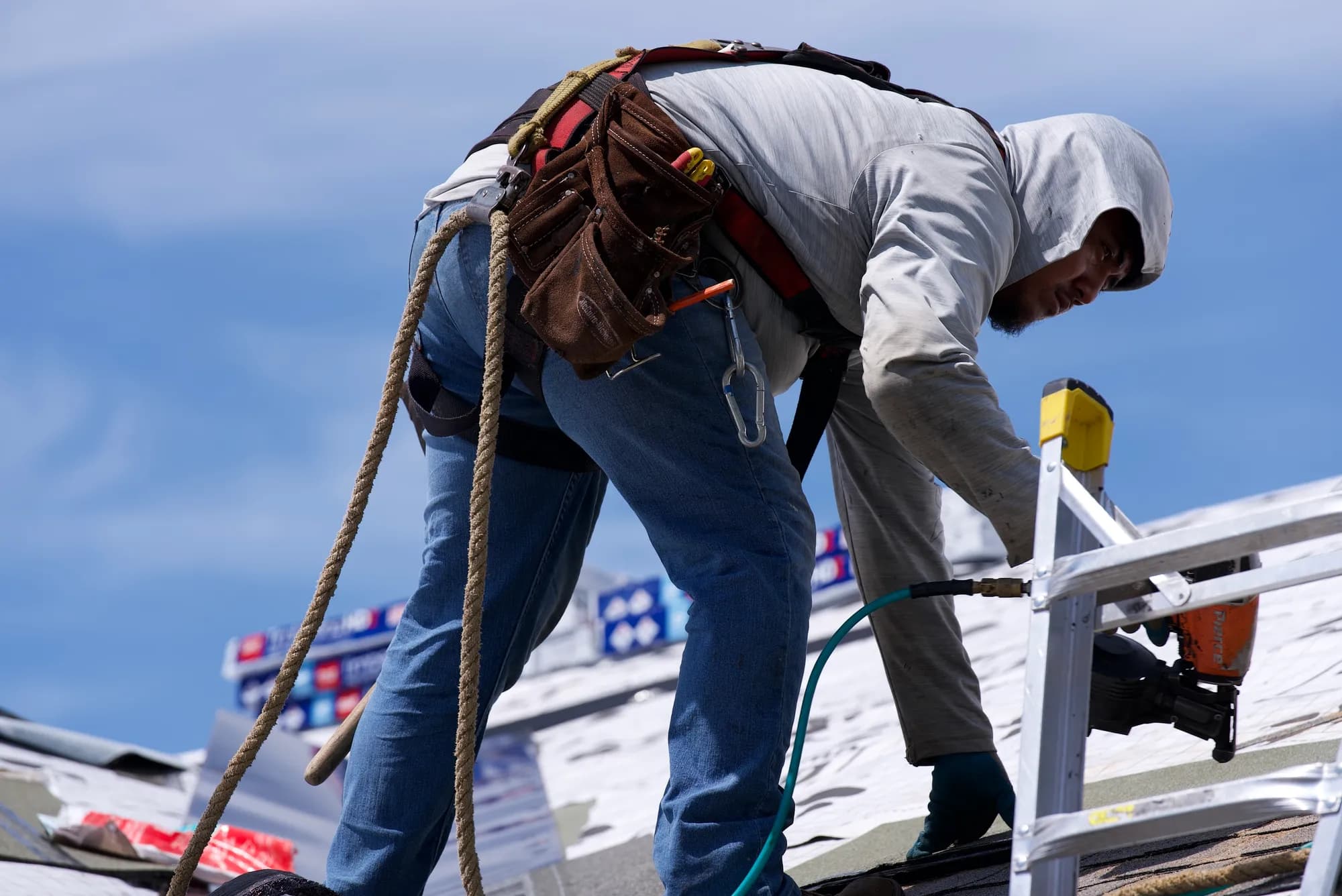 A harnessed roofer handling a pneumatic nail gun and air hose on underlayment in Hillsboro, OR