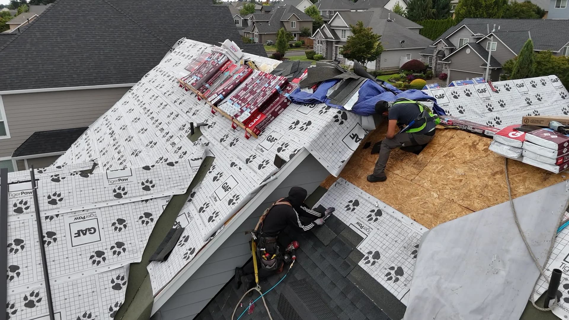 Two harnessed roofers working among GAF Tiger Paw underlayment, exposed OSB sheathing, and staged shingle bundles in Salem, OR