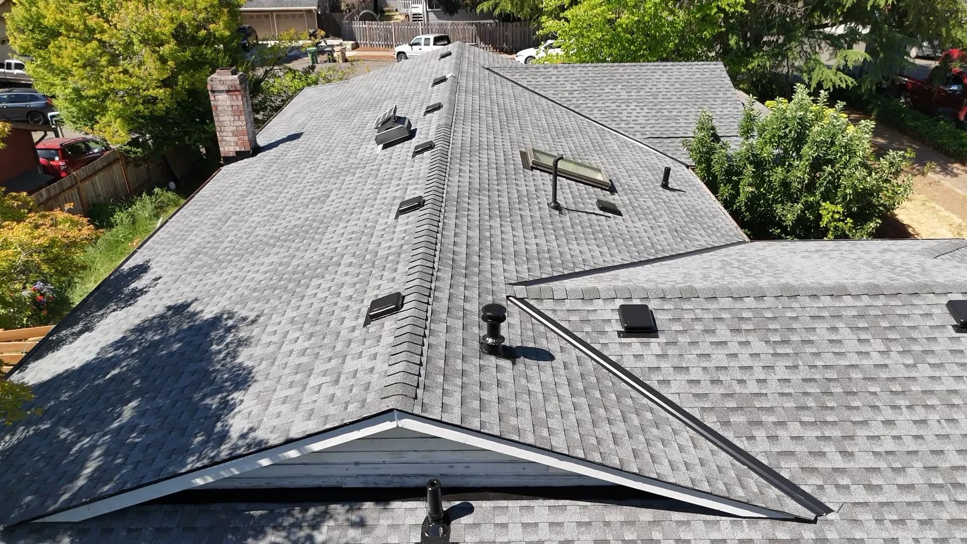 Low aerial view along the ridge of a completed gray shingle roof with a skylight and brick chimney in Tualatin, OR