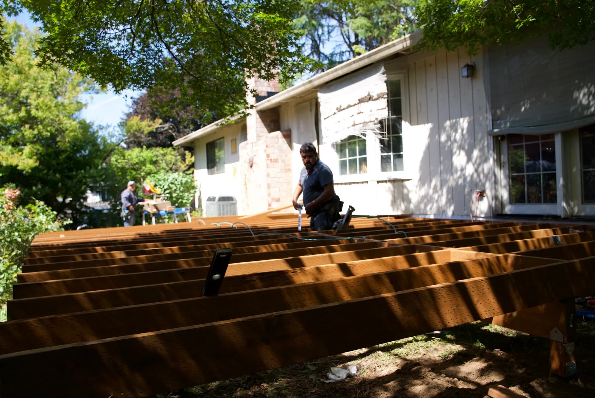 A worker kneeling on parallel wooden deck joists while another cuts lumber at a miter saw in Tualatin, OR
